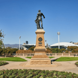 A bronze statue of a man stands on a tall stone plinth in a landscaped park, with circular flower beds in the foreground and modern stadium structures in the background under a clear blue sky.