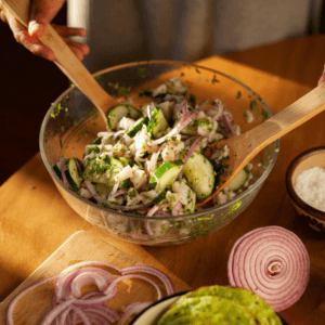 A person mixes a fresh salad with cucumber, red onion, and herbs in a glass bowl using wooden utensils. Sliced red onion and a bowl of salt are on the wooden table nearby.