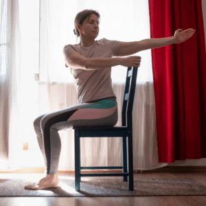 A person sitting on a chair indoors stretches their right arm across their chest, holding the chair back with their left hand. Sunlight filters through sheer curtains and red drapes hang to the side.