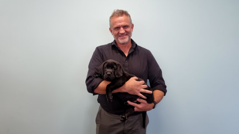 A smiling man in a dark shirt stands against a plain light blue wall, holding a small black Labrador puppy in his arms.