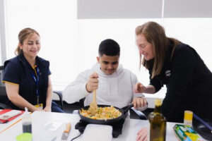 A young person stirs vegetables in a pan with a wooden spoon, while two women sit and stand beside him, smiling and observing at a white table with cooking items.