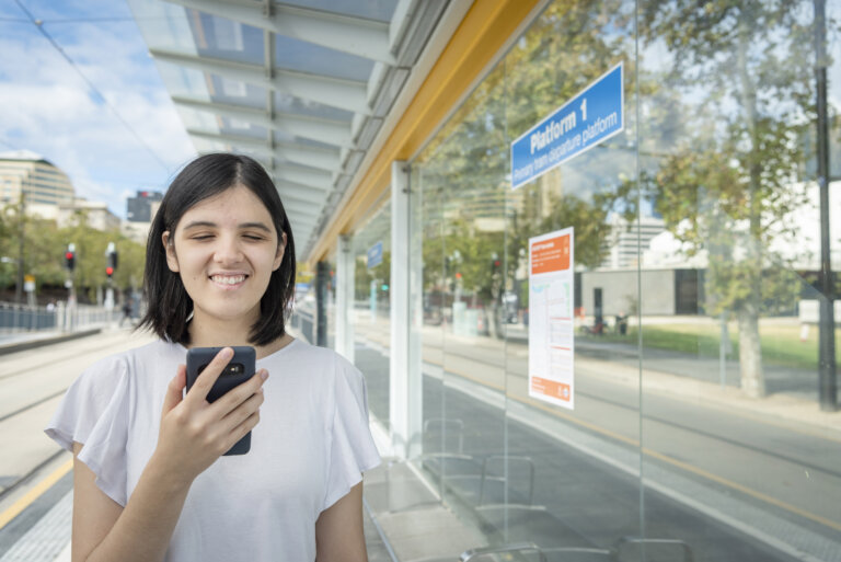 A young woman with red hair and a white lace dress smiles whilst holding a mobile phone with a decorative case. Blurred flowers and people are in the bright background.