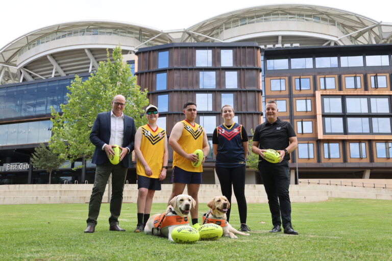 Five people, three in yellow or navy sports kits, stand on grass with two guide dogs and fluorescent yellow footballs in front of a modern stadium and building. The scene appears to promote an inclusive sporting event.