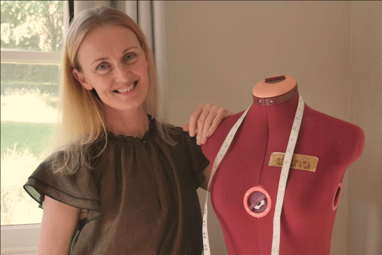 A smiling woman with blonde hair stands indoors next to a red dressmaker’s dummy draped with a measuring tape. Sunlight streams through a window in the background.