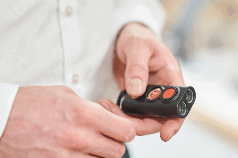 A person wearing a white shirt holds a small black remote control device with red and orange buttons, using their thumb to press one of the buttons.