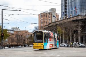 A yellow and white tram with colourful adverts travels through a city intersection, surrounded by tall buildings, traffic lights, and a few pedestrians on a cloudy day.