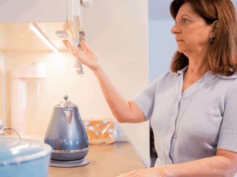 A woman standing in a kitchen turns on an under-cabinet light with a wave of her hand. A blue electric kettle sits on the worktop in front of her.