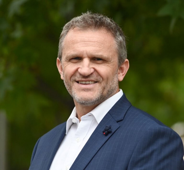 A middle-aged man with short, light brown hair and a trimmed beard is smiling. He is wearing a dark blue suit and white shirt, standing outdoors with lush green foliage in the background.