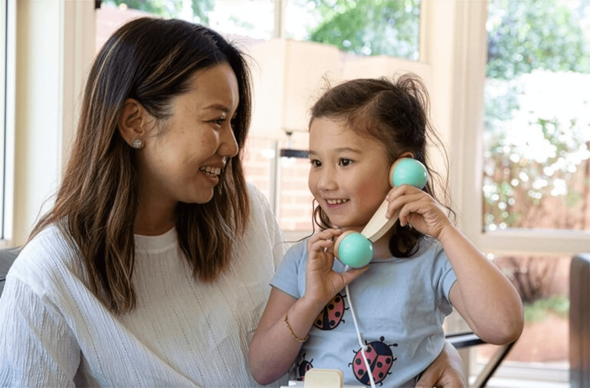 A woman smiles at a young girl who is holding a toy telephone to her ear. The girl wears a blue shirt with ladybirds, and they are sitting together in a cheerful space filled with sunlight from large windows.