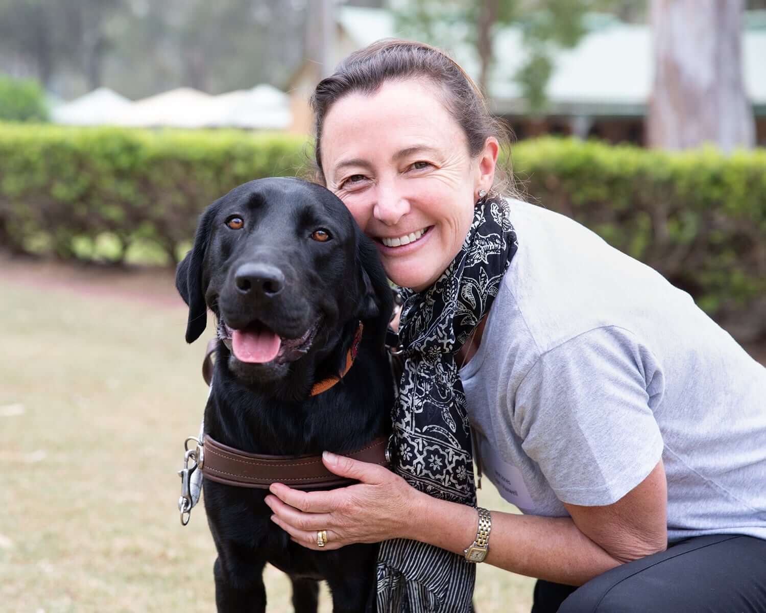 A smiling woman kneels next to a black Labrador in a harness, outdoors on a grassy area. She hugs the assistance dog, and both look happy. Bushes and trees are in the background.