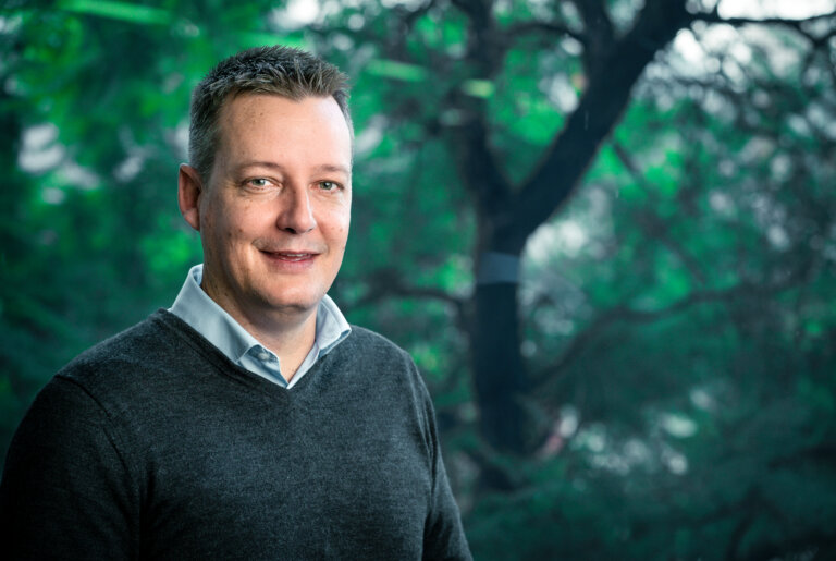A man with short brown hair, wearing a grey jumper over a light blue collared shirt, smiles at the camera. He stands in front of a window with green trees and leafy branches visible outside.