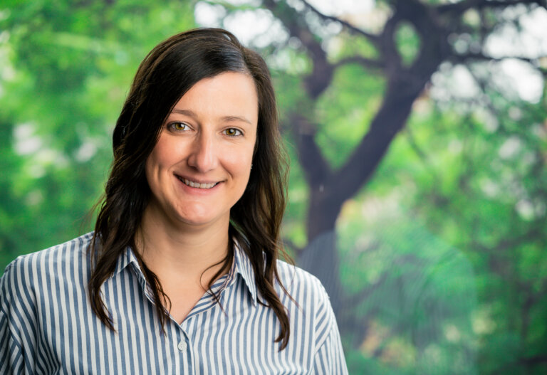 A woman with long brown hair, wearing a blue and white striped shirt, smiles at the camera. The sunlit background is blurred and features green trees.