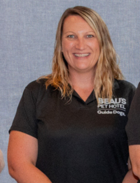 A smiling woman with wavy blonde hair stands in front of a grey background, wearing a black polo shirt embroidered with BEAUS PET HOTEL Guide Dogs.