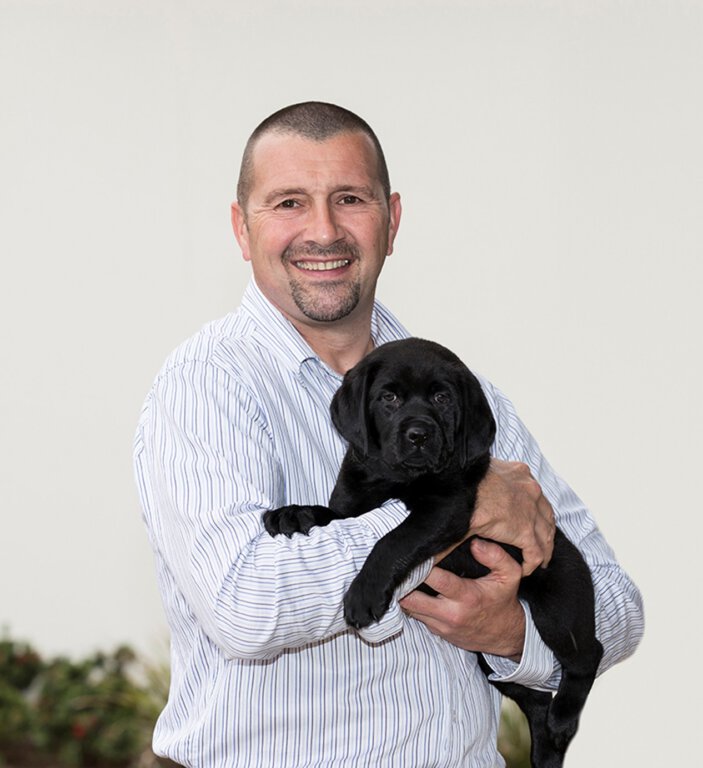 A smiling man in a striped shirt holds a playful black Labrador puppy in his arms, standing outdoors against a light background with lush greenery visible below.