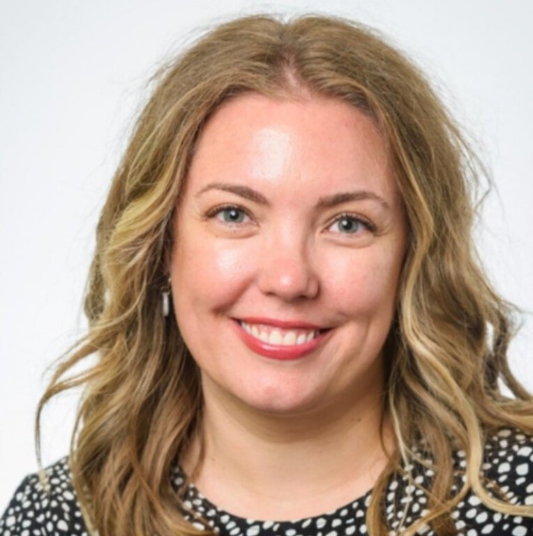 A woman with wavy blonde hair, light skin, and blue eyes smiles at the camera. She is wearing a black and white polka dot top against a light background. Her cheerful expression enhances the lively atmosphere of the image.