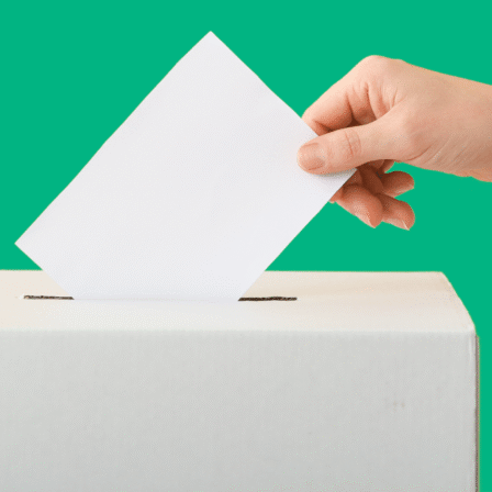 A hand is placing a white ballot into a slot on top of a white ballot box, set against a solid green background.