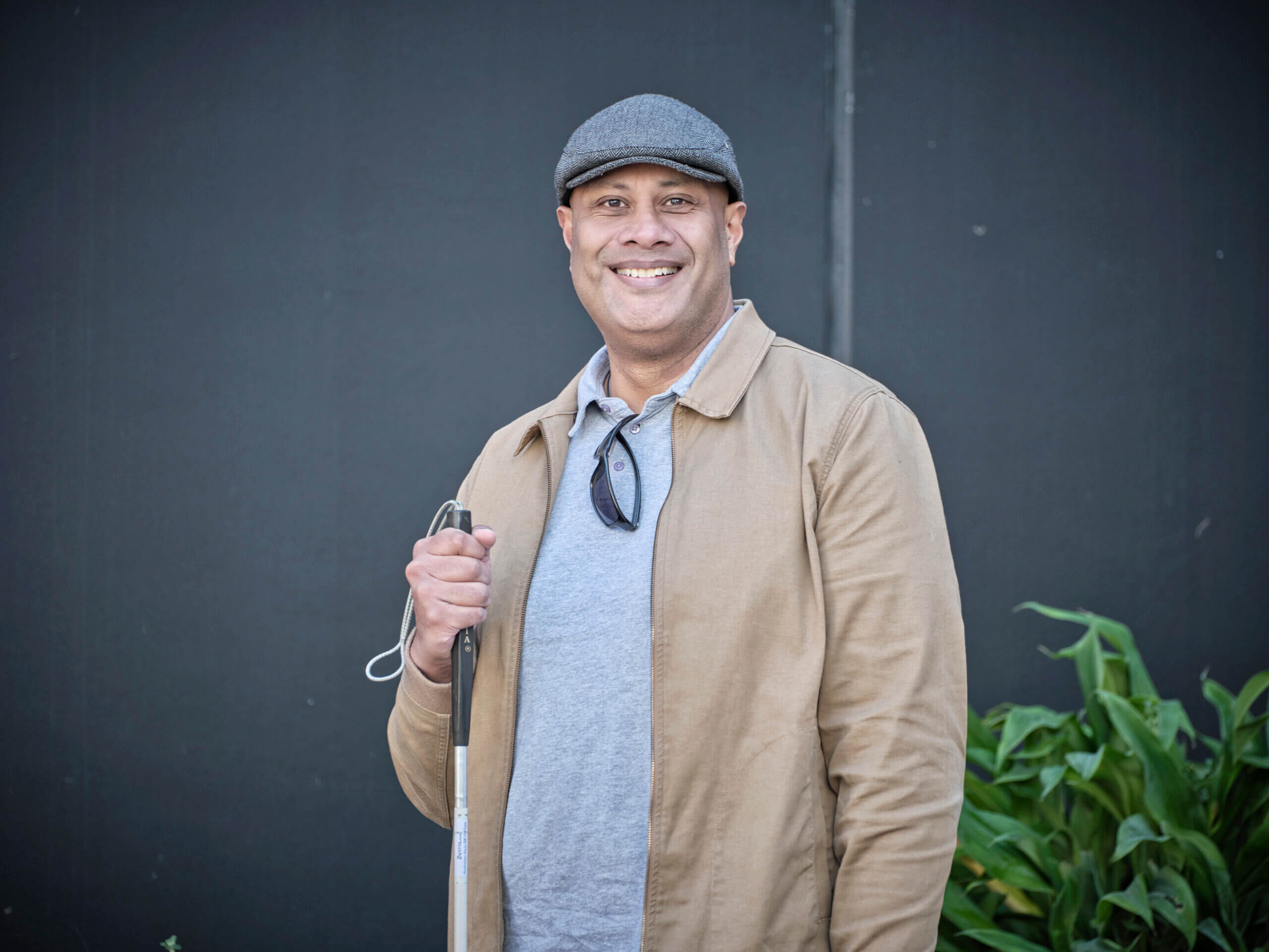 A smiling man in a grey flat cap and beige jacket stands in front of a dark wall, holding a white cane. Green plants are visible to the right side of the image.