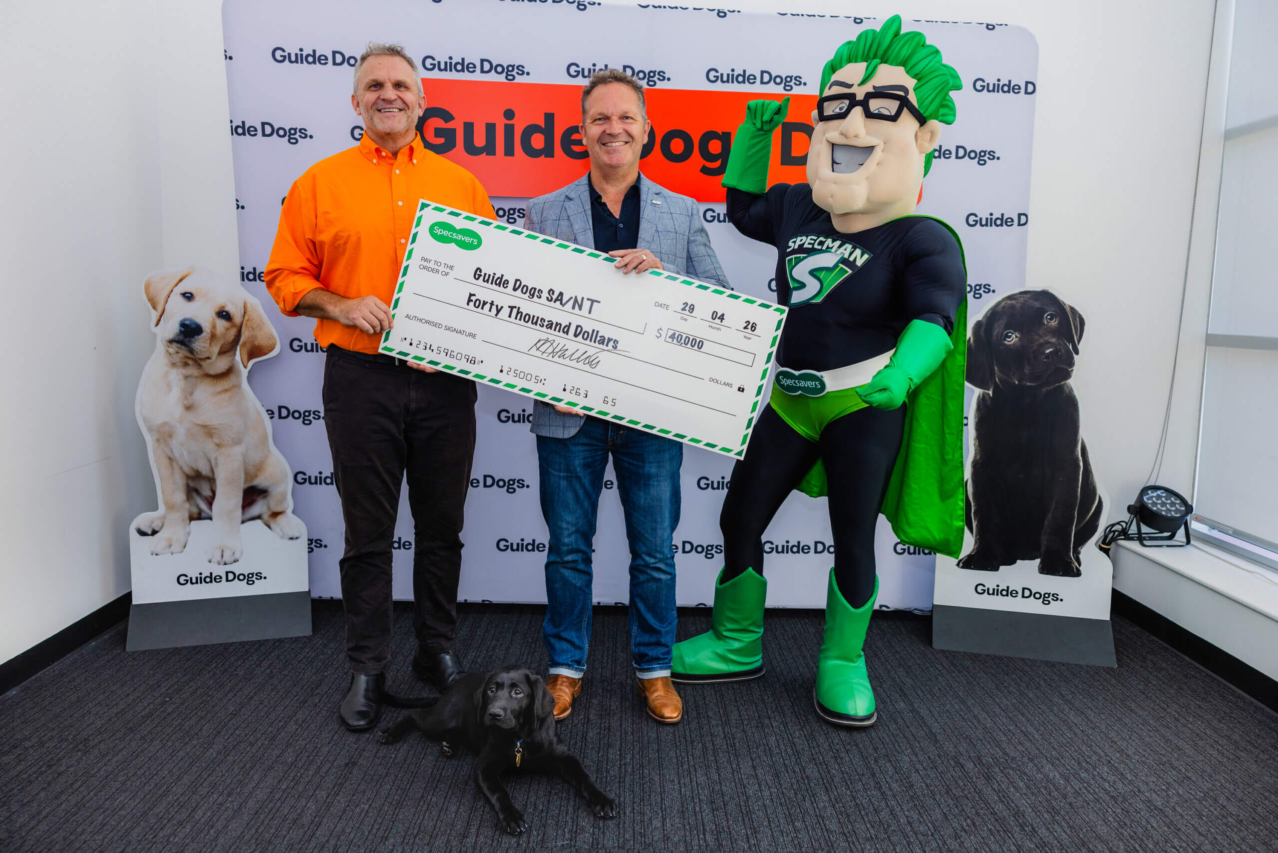 Two smiling men hold a large cheque for £40,000 made out to Guide Dogs SA/NT, standing with a black Labrador and a person in a green superhero costume. Guide Dogs banners and dog images are in the background.