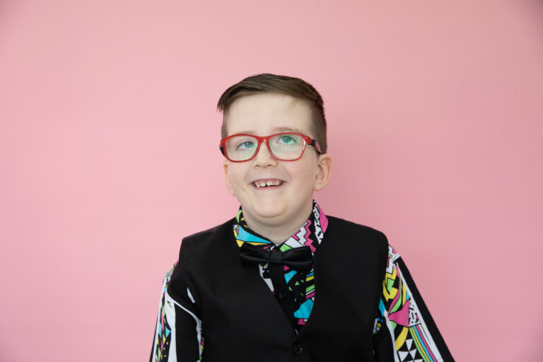 A smiling boy with short brown hair and red glasses wears a colourful patterned shirt, black waistcoat, and bow tie. He poses in front of a solid pink background.