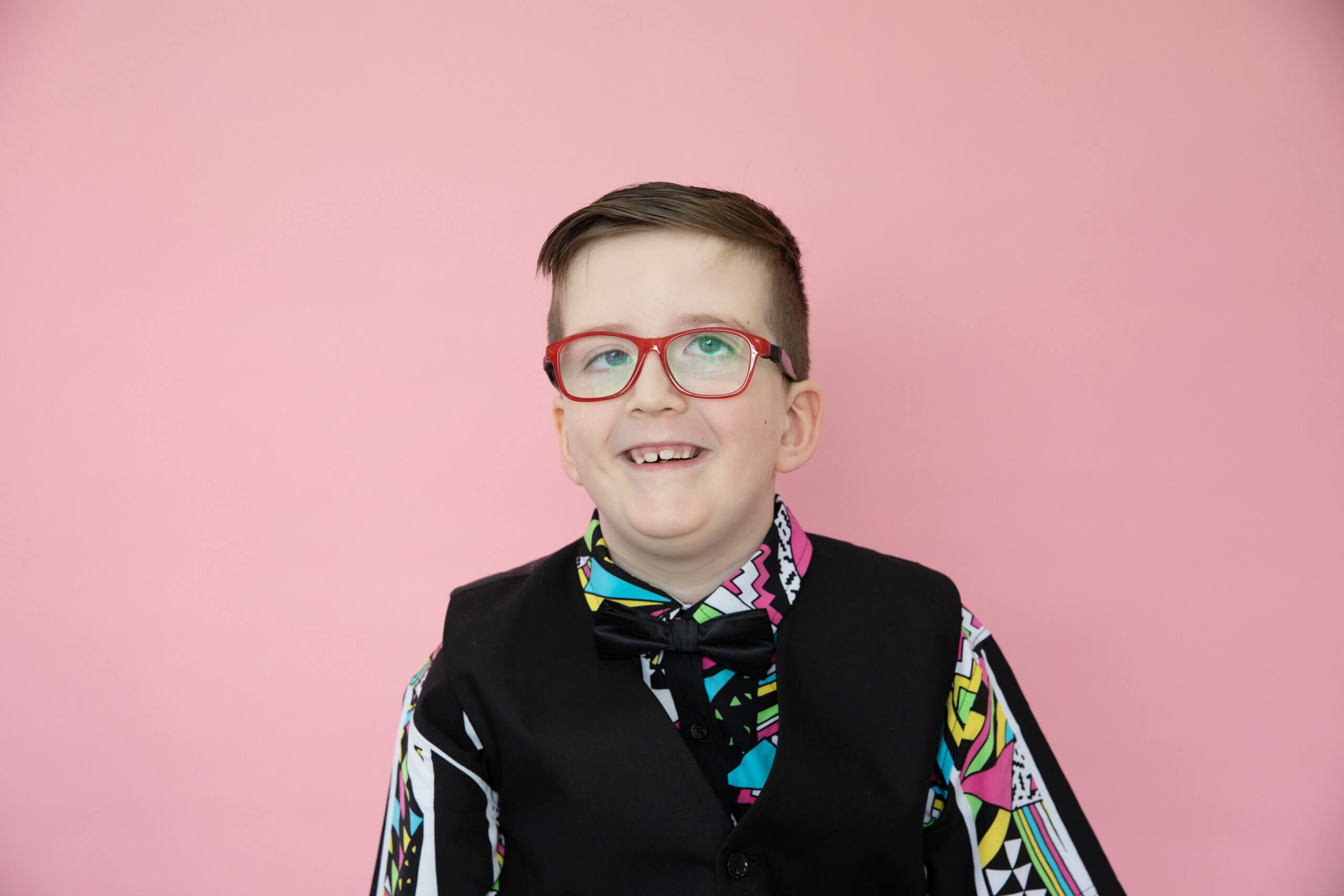 A smiling boy with short brown hair and red glasses wears a colourful patterned shirt, black waistcoat, and bow tie. He poses in front of a solid pink background.