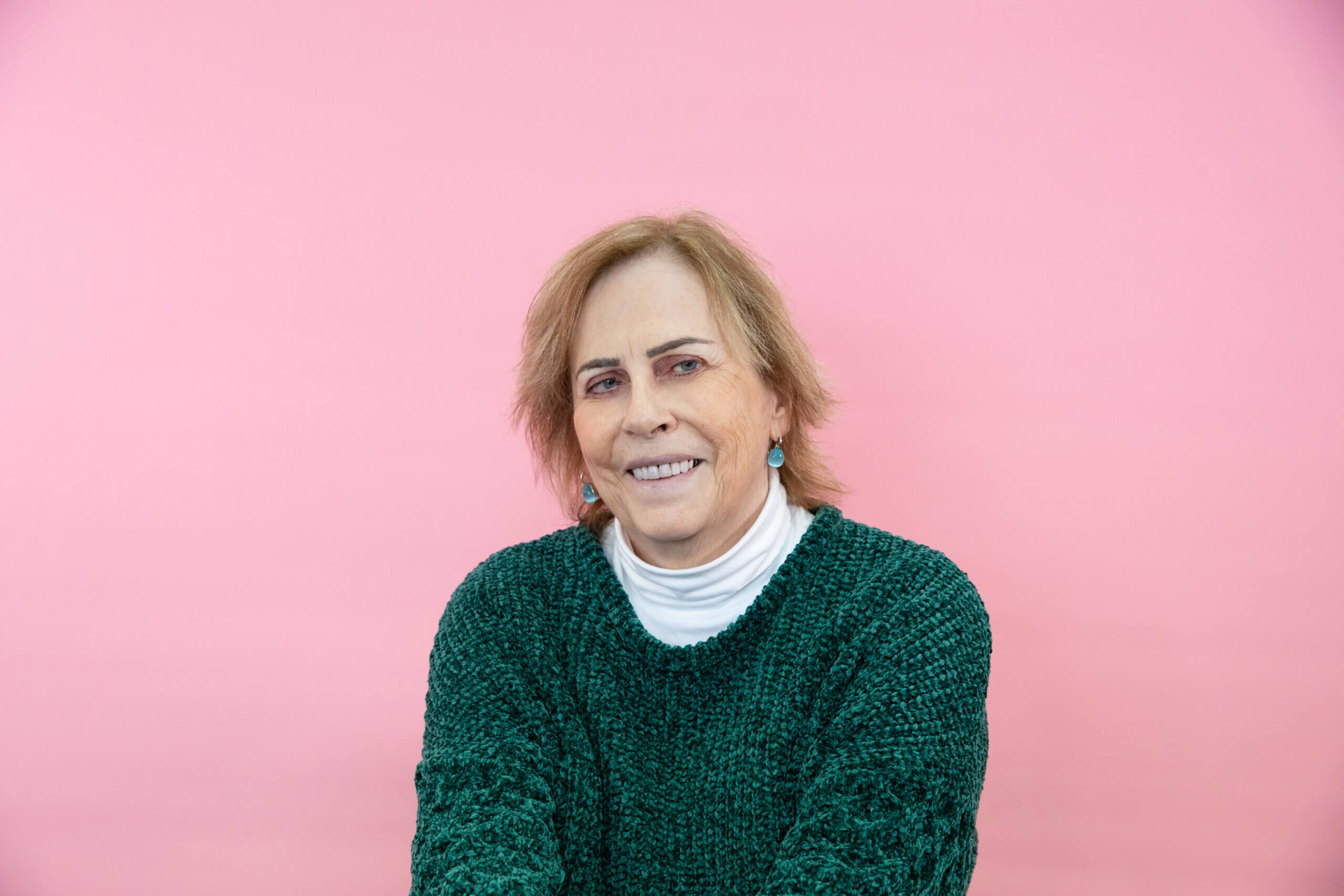 A person with short light brown hair, wearing a cosy green knitted jumper over a white polo neck, smiles cheerfully in front of a solid pink background.