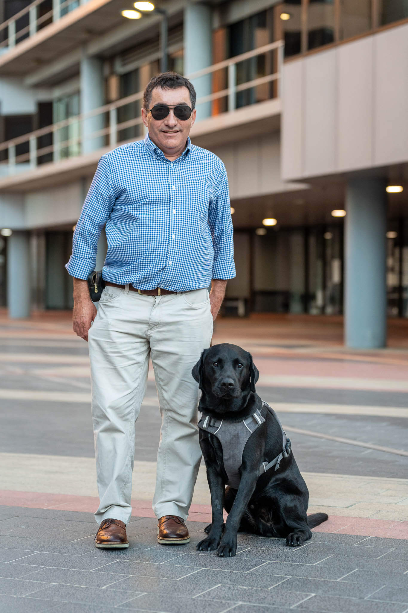 A man wearing sunglasses, a blue checked shirt, and beige trousers stands outside a modern building with a black guide dog sitting by his side, both looking towards the camera.