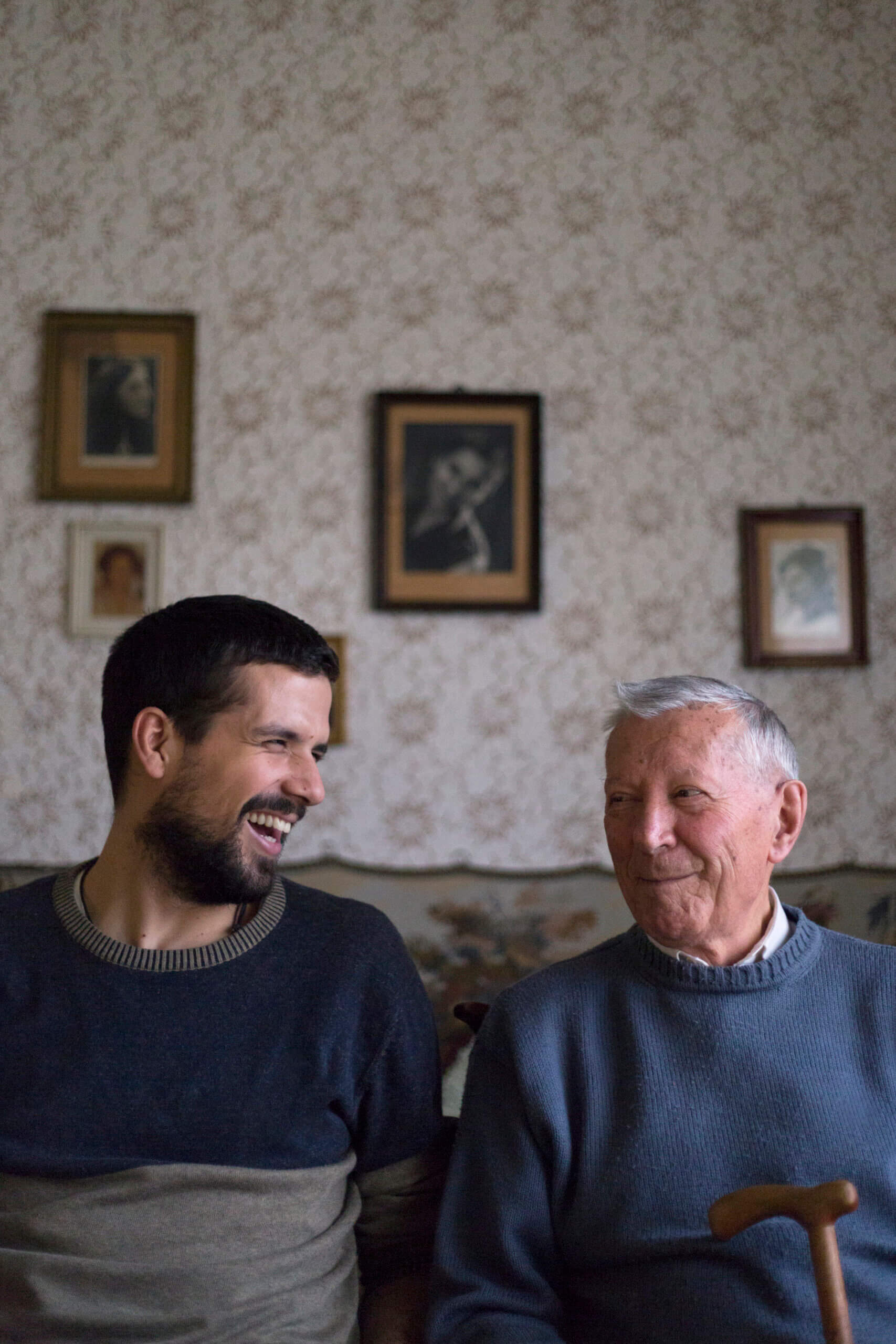 A young man and an elderly man sit side by side on a sofa, smiling and looking at each other. Behind them, framed photos hang on a patterned wall. The elderly man holds a walking stick.