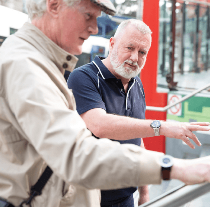 Two older men are standing outdoors. One, wearing a light jacket and hat, is in the foreground pointing at something, whilst the other, in a dark polo shirt, looks on attentively, gesturing with his hand.