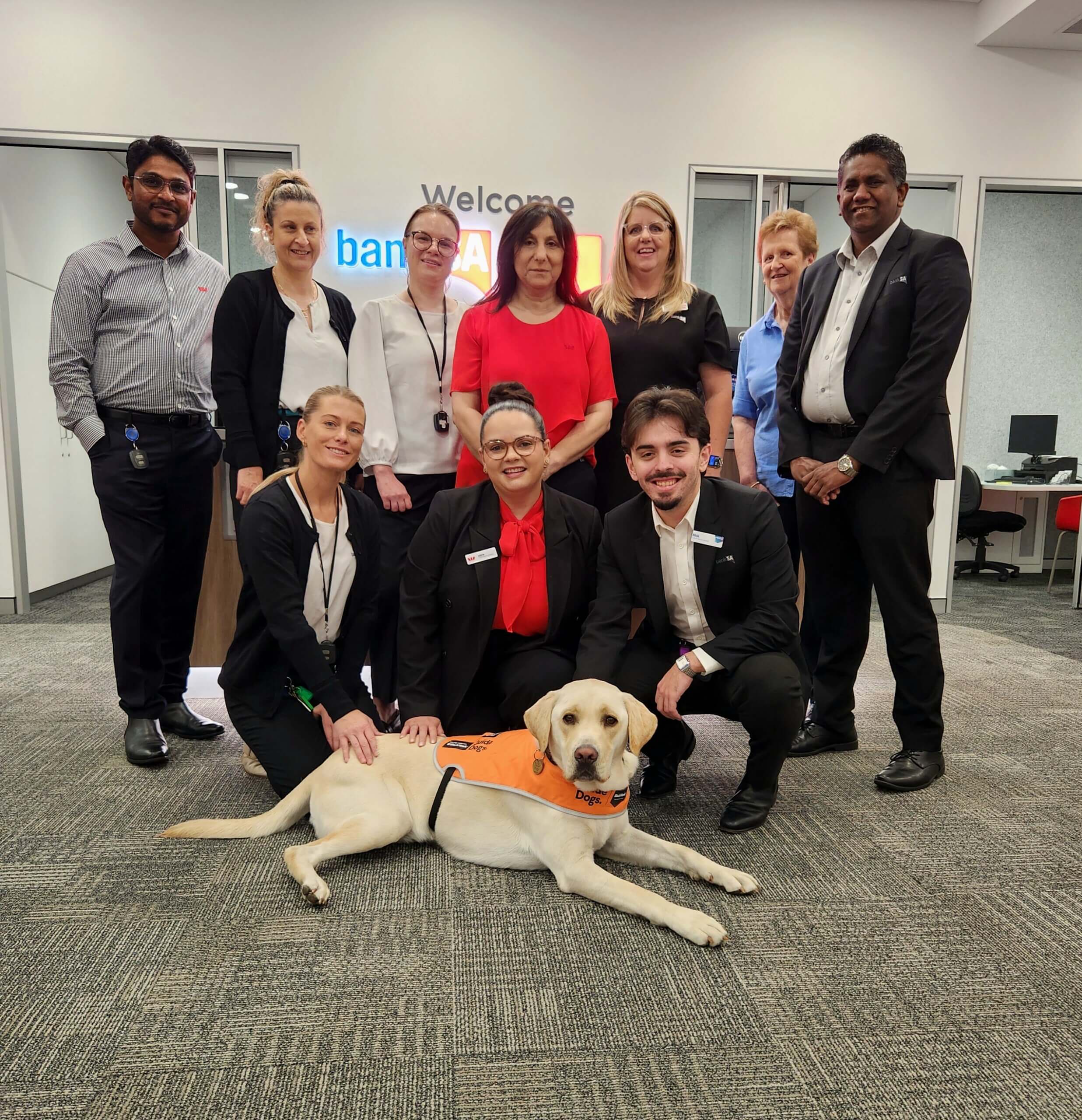 A group of eleven people, some standing and some kneeling, pose and smile inside a bank office. In front, a yellow guide dog wearing an orange vest lies on the carpeted floor. The bank's logo is visible on the wall behind them.