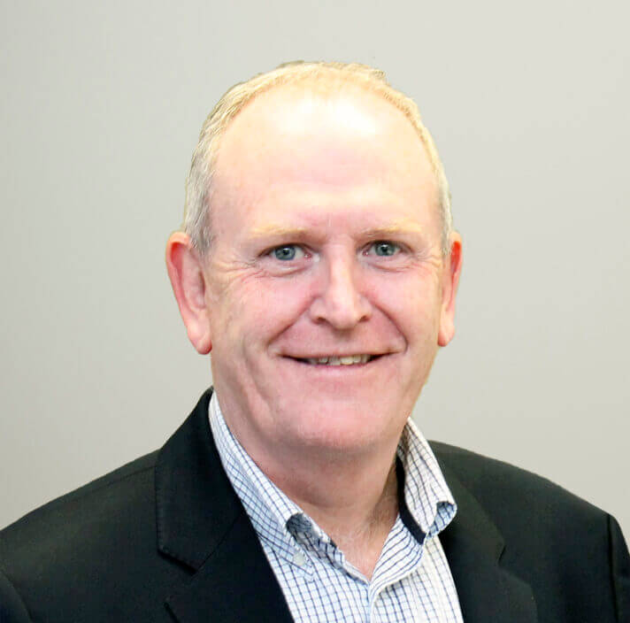 A middle-aged man with short, fair hair is smiling warmly. He is wearing a dark jacket over a checked shirt and is posed confidently in front of a plain, light grey background.