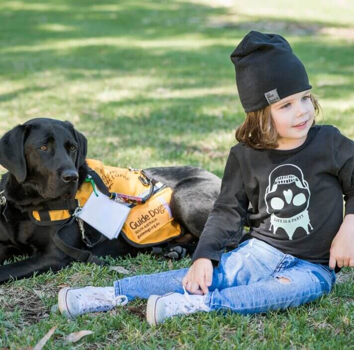 A young child in a black beanie and ripped jeans sits on grass next to a black guide dog wearing a yellow vest. Both are outdoors on a sunny day, and the child smiles whilst looking away.