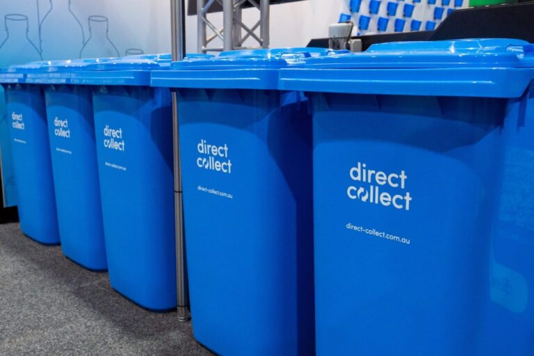 Row of blue recycling bins with the label direct collect printed on the front, lined up indoors on a grey floor with a blurred background.
