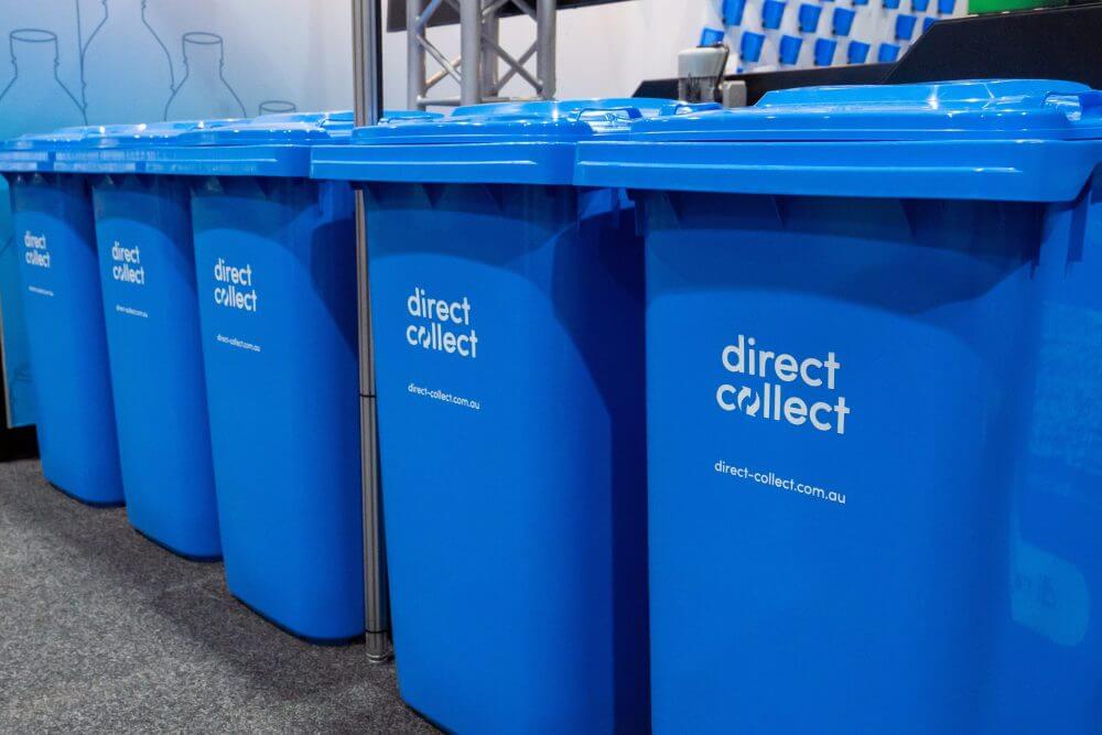 Row of blue recycling bins with the label direct collect printed on the front, lined up indoors on a grey floor with a blurred background.