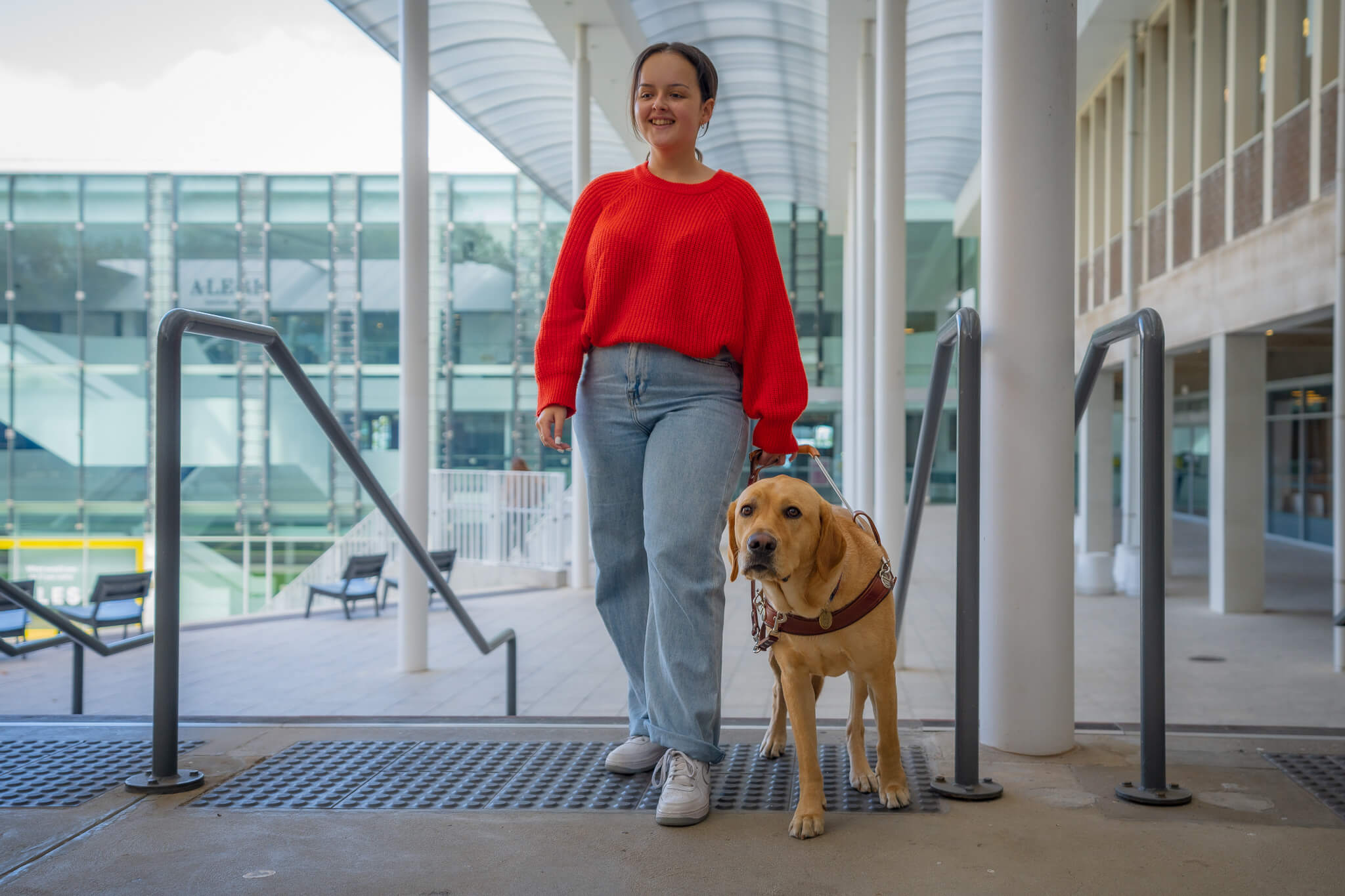 A woman in a red jumper and jeans walks down a ramp while holding the harness of a guide dog. The background shows a modern glass building and outdoor seating.