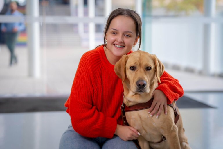 A smiling young woman in a bright red jumper sits indoors, hugging a yellow Labrador guide dog wearing a harness. They are both looking forward, sitting on a shiny floor with a blurred background.
