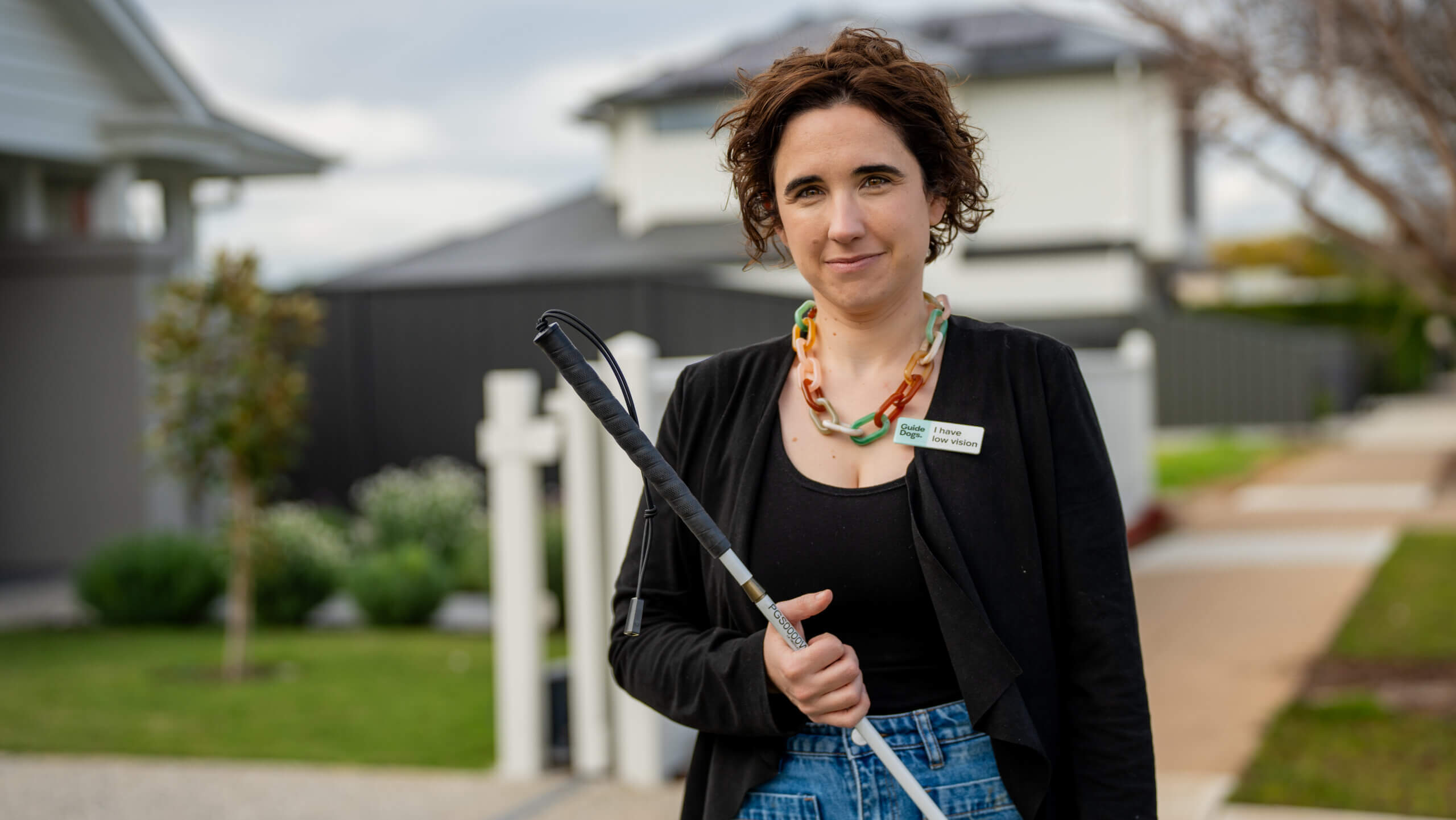 A woman with curly brown hair stands outdoors in a neighbourhood, holding a white cane. She wears a black jacket, patterned necklace, and blue jeans. A name tag is pinned to her jacket as she stands near houses in the background.