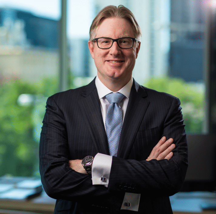 A smiling man in a dark pinstripe suit and light blue tie stands confidently with arms crossed in a modern office with large windows and a blurred cityscape in the background.