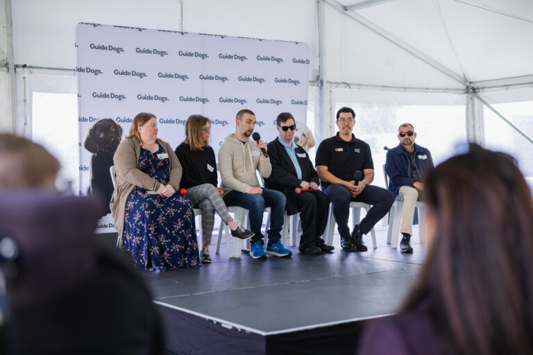 Six people sit on a stage under a marquee, participating in a panel discussion. A Guide Dogs banner is behind them. The audience is visible in the foreground.