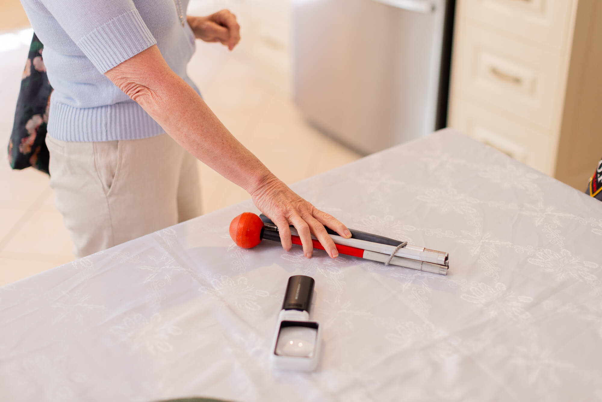 A person’s hand reaches for metal tongs with red silicone tips on a white tablecloth, whilst a black and white magnifying glass rests nearby. A kitchen setting is visible in the background.