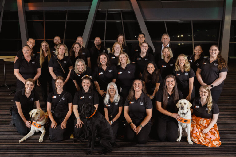 A group of 26 adults, mostly women in matching black shirts, pose indoors for a group photo. Four guide dogs—three yellow and one black—sit at the front as some group members smile behind them, celebrating teamwork and support.