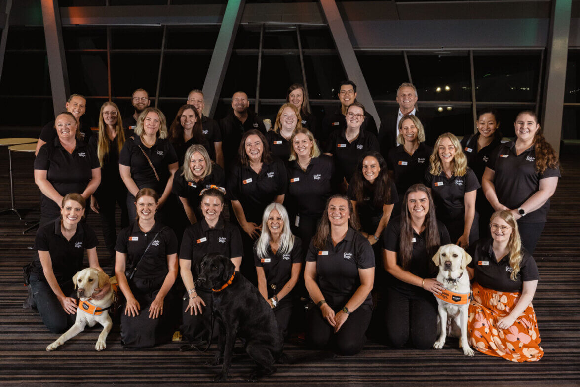 A group of 26 adults, mostly women in matching black shirts, pose indoors for a group photo. Four guide dogs—three yellow and one black—sit at the front as some group members smile behind them, celebrating teamwork and support.