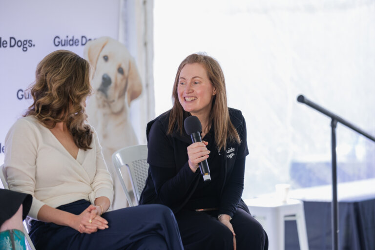 Two women sit and talk on stage; one holds a microphone and smiles. Behind them is a banner with Guide Dogs and an image of a yellow Labrador Retriever. A microphone stand is visible to the right.