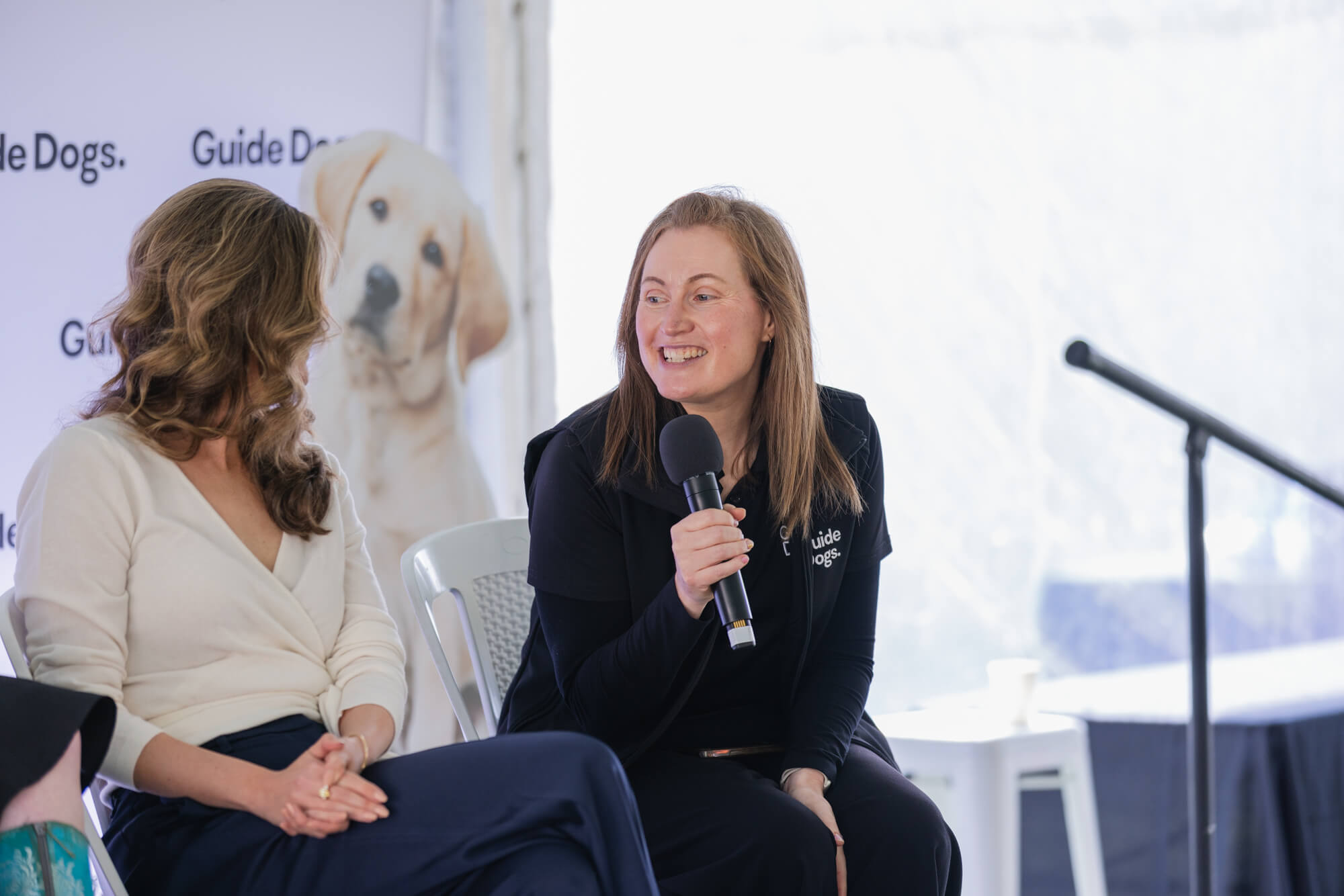 Two women sit and talk on stage; one holds a microphone and smiles. Behind them is a banner with Guide Dogs and an image of a yellow Labrador Retriever. A microphone stand is visible to the right.