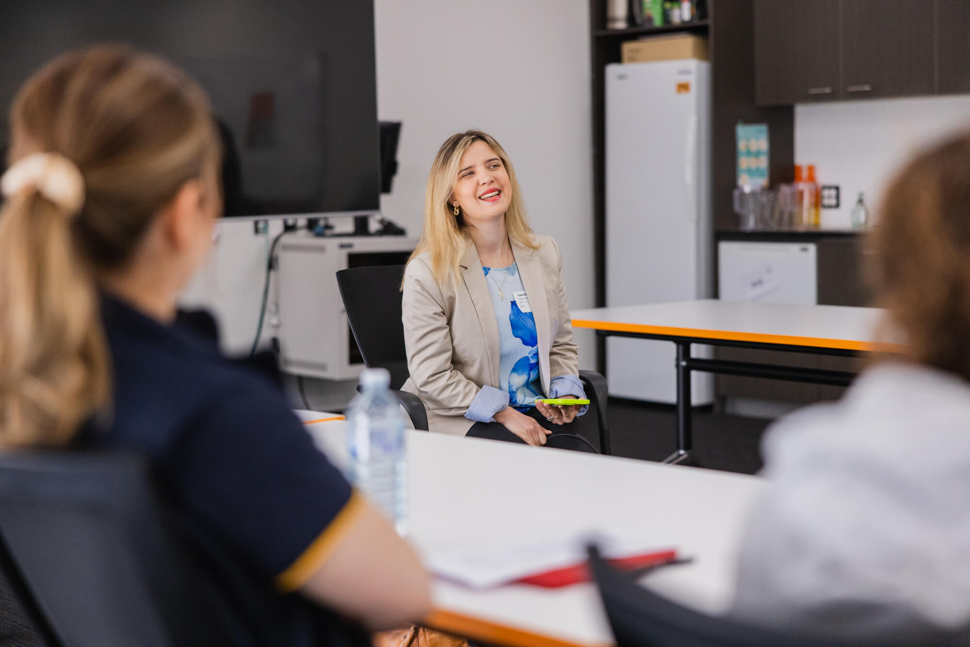 A woman with blonde hair and a beige blazer sits smiling and talking to two people in an office or classroom setting, with tables, chairs, and a water bottle visible.