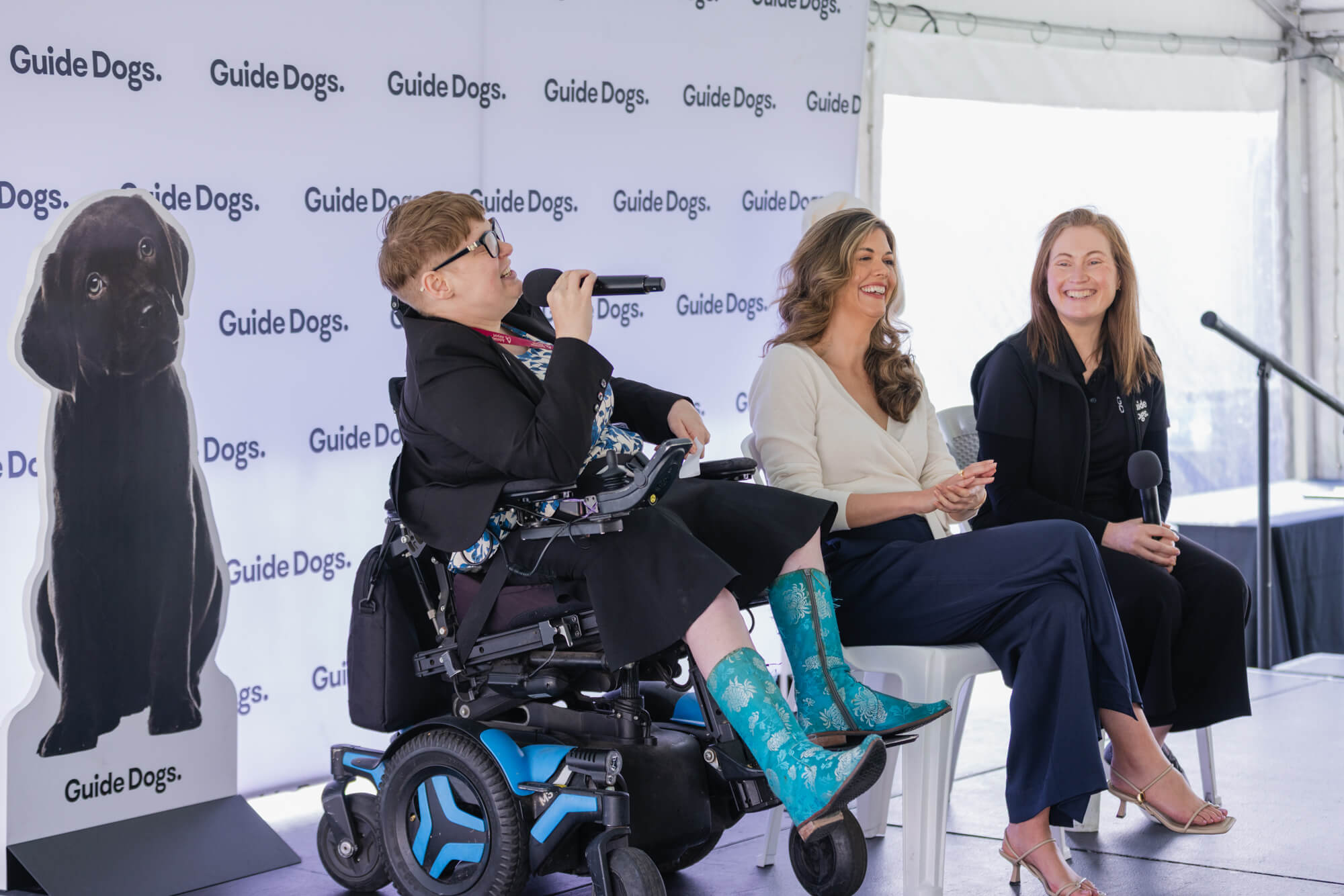 Three women sit on a stage, one in a wheelchair holding a microphone, with Guide Dogs branding behind them and a large dog photo cut-out. Two women are smiling, one holding a microphone, at an outdoor event.