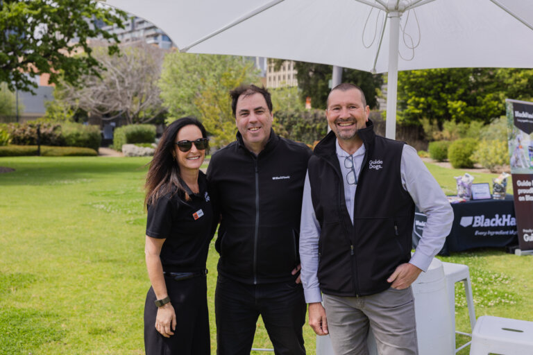 Three adults stand and smile outdoors under a white umbrella at an event. Two men wear black jackets, and a woman wears a black shirt and sunglasses. Tables and banners are visible in the background on a grassy lawn.