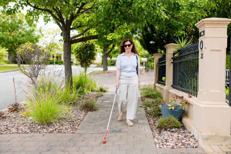 A woman with long brown hair, wearing sunglasses and light-coloured clothes, walks on a pavement using a white cane. She is surrounded by greenery and residential fencing on a sunny day.