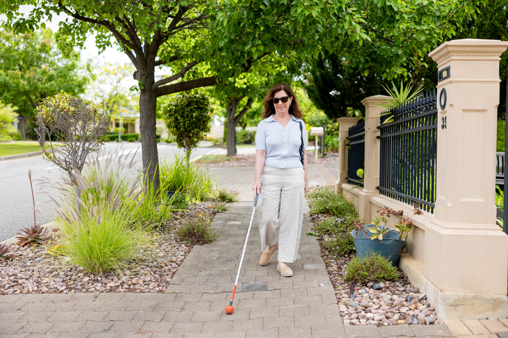 A woman with long brown hair, wearing sunglasses and light-coloured clothes, walks on a pavement using a white cane. She is surrounded by greenery and residential fencing on a sunny day.