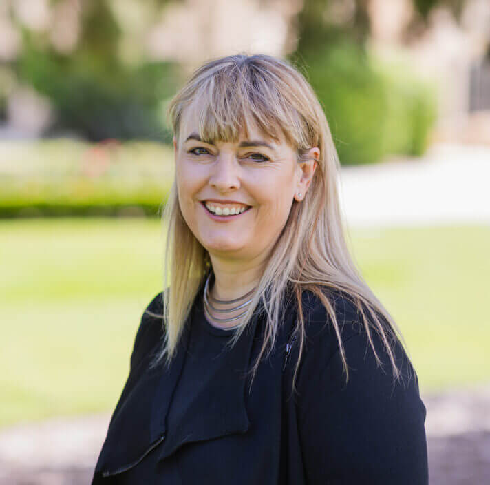 A woman with long blonde hair and fringe smiles outdoors, wearing a black jacket and layered necklace, with a blurred green lawn and trees in the background, exuding confidence.