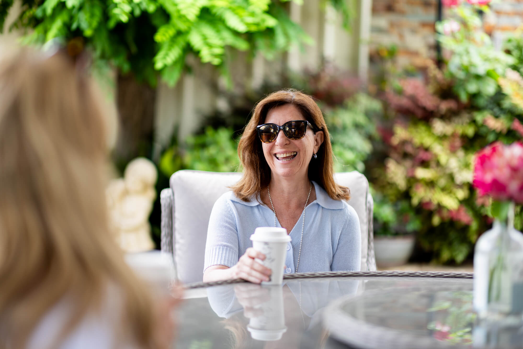 A woman in sunglasses and a light blue shirt sits at an outdoor glass table, smiling and holding a takeaway coffee cup. Surrounded by lush greenery and flowers, she enjoys her coffee whilst another person is blurred in the foreground.